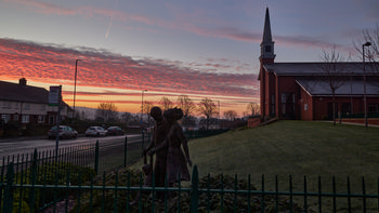 Chesterfield church sunrise This landscape photograph shows a winter sunrise at the Church of Jesus Christ of the Latter Day Saints in Chesterfield, Derbyshire, United Kingdom. The image captures the church’s modern architecture with its tall steeple, set against an orange and pink morning sky. In the foreground, a statue depicting a family stands next to a street lined with parked cars and traditional British houses. Leafless trees are visible on and around the church grounds, further indicating the season. The sunlight highlights the silhouettes of the church and trees, providing a striking view of this Chesterfield location.
